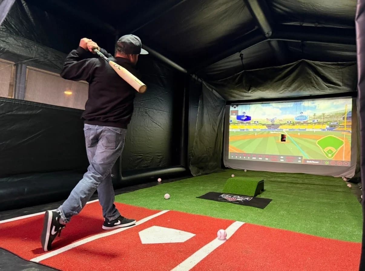 A player mid-swing inside the On Deck hit house, the ball tracked across a big-league stadium on the screen